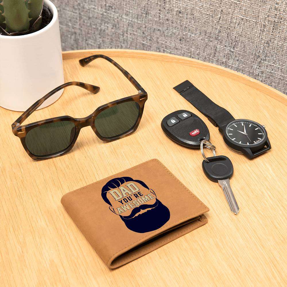 Leather wallet with "Dad You're Awesome" design on wooden table surrounded by sunglasses, watch, and keys.